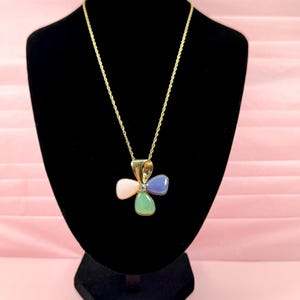 May include: A gold-tone rope chain necklace featuring a pendant shaped like a four-leaf clover. The pendant consists of four smooth, teardrop-shaped stones in pastel pink, light blue, and green, with a small clear stone set in the centre.