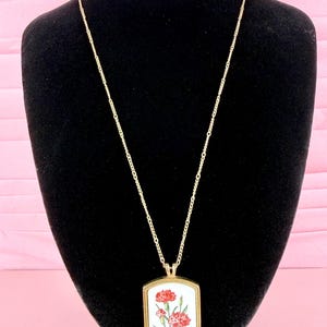May include: A gold-tone necklace featuring a rectangular pendant with a white background and a detailed illustration of two red carnation flowers with green stems and leaves. The pendant is suspended from a delicate gold-tone link chain.