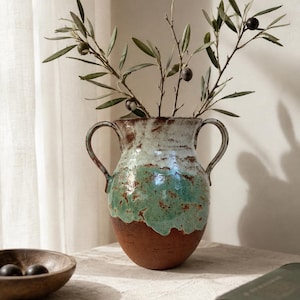 Könnte beinhalten: Eine Keramikvase mit zwei Henkeln im rustikalen Design mit terrakottafarbenem Boden, türkisfarbener Tropfglasur und weißem Oberteil. Die Vase enthält einen Zweig mit grünen Blättern und kleinen dunklen Oliven mit einer strukturierten Oberfläche.