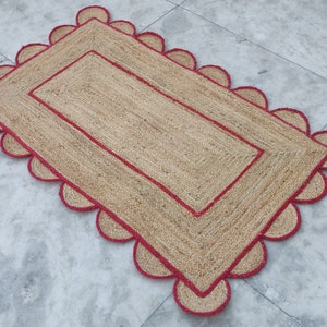 Puede incluir: Una alfombra rectangular de yute con un borde rojo y bordes festoneados. La alfombra tiene una textura tejida y un color marrón natural.