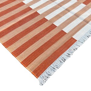 K&ouml;nnte beinhalten: Nahaufnahme eines gestreiften Teppichs mit wei&szlig;em Fransen. Der Teppich hat abwechselnde Streifen in Orange und Beige.