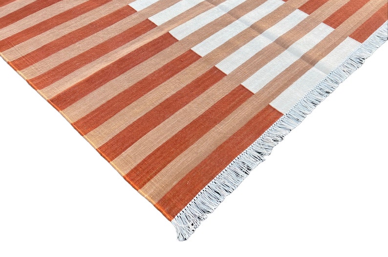 K&ouml;nnte beinhalten: Nahaufnahme eines gestreiften Teppichs mit wei&szlig;em Fransen. Der Teppich hat abwechselnde Streifen in Orange und Beige.