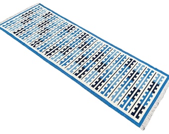 Op de afbeelding: Een blauwe en witte geometrisch gepatroneerde loper met franjes aan de uiteinden. De loper heeft een witte achtergrond met blauwe en zwarte geometrische vormen.