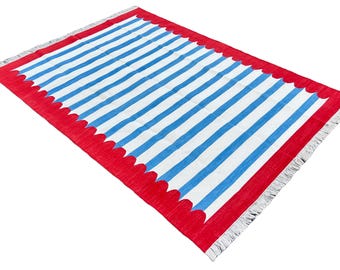 Op de afbeelding: Een rechthoekig vloerkleed met blauwe en witte strepen. Het heeft een rode rand met een geschulpte rand en franjes aan de uiteinden. Het vloerkleed is geschikt voor binnen of buiten. De afmetingen van het vloerkleed zijn in centimeters.