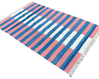Op de afbeelding: Een gestreept tapijt met horizontale roze, blauwe en witte strepen. Het tapijt heeft een franje rand.