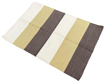 Op de afbeelding: Een rechthoekig textiel met verticale strepen in wit, beige en donkerbruin. De stof heeft een geweven textuur. De strepen zijn gelijkmatig verdeeld over het textiel, wat een theedoek of een klein tapijt kan zijn.