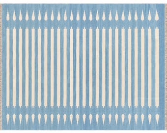 Könnte beinhalten: Ein rechteckiger Teppich mit hellblauem Hintergrund und vertikalen cremefarbenen Streifen. Das Design zeigt ein sich wiederholendes Muster aus kleinen, spitzen, blattartigen Formen an den oberen und unteren Rändern sowie kurze beige Fransen.