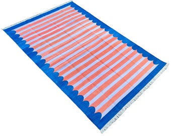 Op de afbeelding: Een roze en wit gestreept tapijt met een blauwe gegolfde rand. Het tapijt heeft een franje rand.