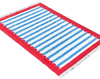 Op de afbeelding: Een rechthoekig tapijt met een streepjespatroon. Het tapijt heeft afwisselend blauwe en witte strepen over het midden. Een rode rand met een geschulpte rand omlijst het streepjespatroon. Het tapijt heeft franjes aan de korte uiteinden.