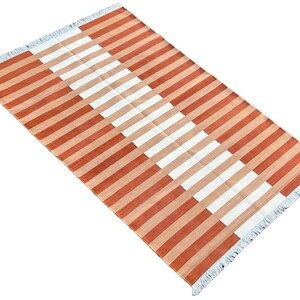K&ouml;nnte beinhalten: Ein rechteckiger Teppich mit einem Streifenmuster in Orange- und Wei&szlig;t&ouml;nen. Der Teppich hat einen Fransenrand.