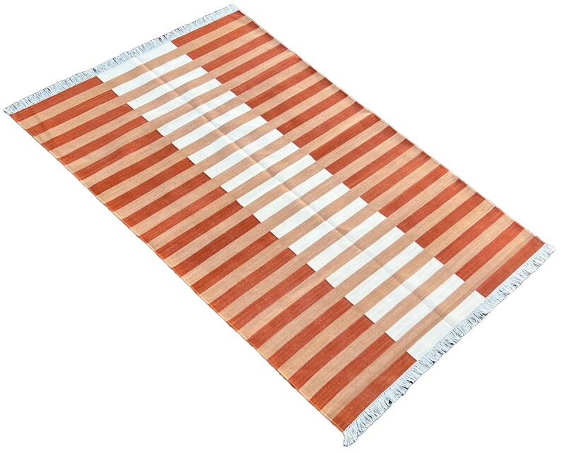 K&ouml;nnte beinhalten: Ein rechteckiger Teppich mit einem Streifenmuster in Orange- und Wei&szlig;t&ouml;nen. Der Teppich hat einen Fransenrand.