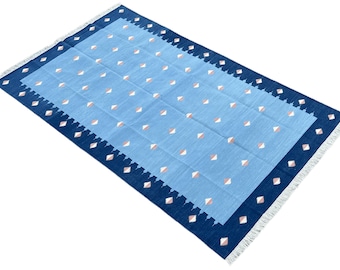 Op de afbeelding: Een blauw en roze gestreept tapijt met een donkerblauwe rand en witte franjes. Het tapijt heeft een geometrisch ontwerp met kleine roze en witte ruiten op een lichtblauwe achtergrond.