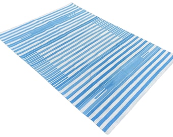 Puede incluir: Una alfombra rectangular con un patrón de rayas en tonos azules y blancos. Las rayas corren en diagonal por la alfombra, con algunas secciones que presentan un diseño de rayas rotas. La alfombra parece estar hecha de un material suave y tejido.