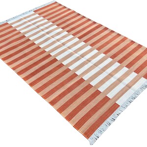 K&ouml;nnte beinhalten: Ein gestreifter Teppich mit einem geometrischen Muster in Orange- und Wei&szlig;t&ouml;nen. Der Teppich hat einen Fransenrand.