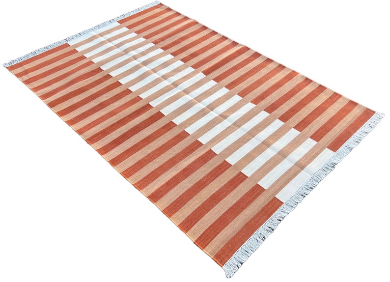 K&ouml;nnte beinhalten: Ein gestreifter Teppich mit einem geometrischen Muster in Orange- und Wei&szlig;t&ouml;nen. Der Teppich hat einen Fransenrand.
