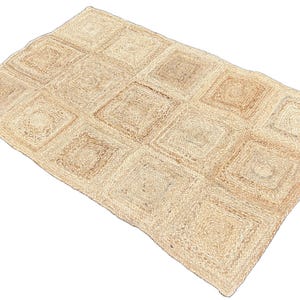 May include: A rectangular rug with a geometric design. The rug is made from natural jute fibres, with a light tan colour. The design features a grid of squares, each with a slightly different texture and shade.
