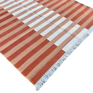 K&ouml;nnte beinhalten: Ein gestreifter Teppich mit einem geometrischen Muster in Orange-, Braun- und Wei&szlig;t&ouml;nen. Der Teppich hat einen Fransenrand.