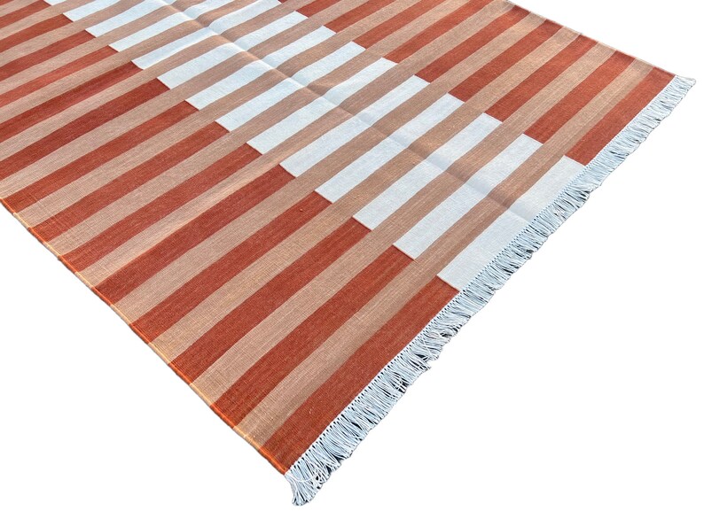 K&ouml;nnte beinhalten: Ein gestreifter Teppich mit einem geometrischen Muster in Orange-, Braun- und Wei&szlig;t&ouml;nen. Der Teppich hat einen Fransenrand.