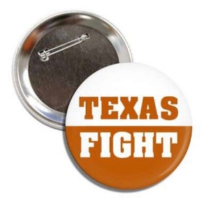 以下が含まれることがあります： 白とオレンジ色の背景に、白いブロック体で「TEXAS FIGHT」と書かれた丸いボタン。ボタンの裏側には金属製のピンが付いています。