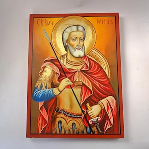 Puede incluir: Icono de un santo, que representa a un hombre con barba blanca y aureola, sosteniendo una lanza y una espada. Viste túnicas rojas y doradas, con una figura con casco a su lado. El fondo es naranja cálido con detalles dorados, y el marco es rojo oscuro.