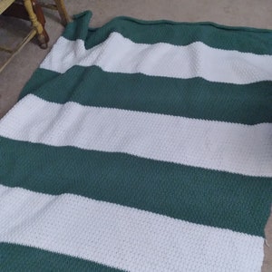 Puede incluir: Una manta de crochet a rayas verdes y blancas. La manta está doblada por la mitad y está sobre una alfombra marrón.