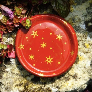 Puede incluir: Un pequeño plato de cerámica rojo con un borde dorado y un patrón de estrellas doradas y puntos.
