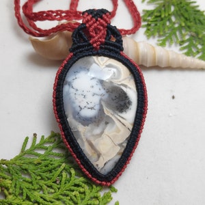 May include: A handmade macrame pendant necklace. The pendant features a teardrop-shaped stone with a black, white, and tan pattern, framed by black and red macrame. The necklace cord is red.