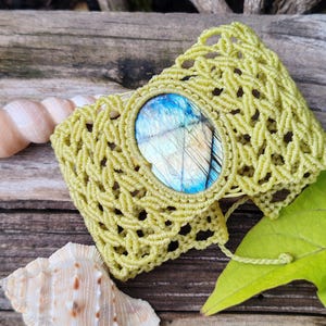 May include: A green macrame bracelet with a blue and green labradorite gemstone. The bracelet is on a wooden surface with a seashell.