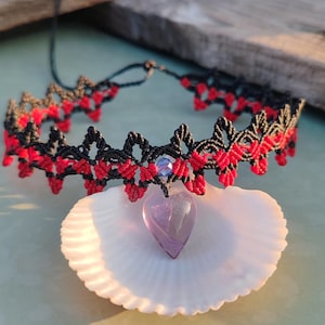 May include: A black macrame choker necklace with red accents and a heart-shaped purple pendant. The necklace is displayed on a white seashell, with a blue gem at the center of the choker.