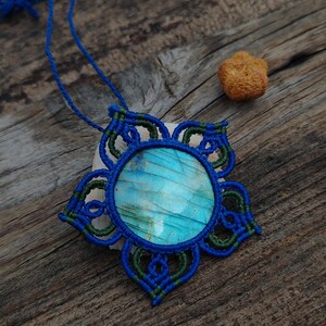 May include: A handcrafted macrame necklace featuring a round, iridescent blue gemstone pendant. The pendant is encased in a blue and green macrame design, with a matching blue cord. The necklace rests on a wooden surface.