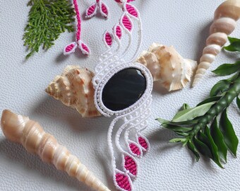 May include: A white and pink macrame necklace with a black oval gemstone pendant. The necklace is displayed on a white surface with seashells and green foliage.