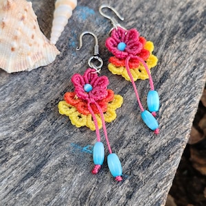 May include: Handmade floral earrings with a crochet design. The earrings feature a vibrant colour palette of red, yellow, and orange, with blue beads dangling from pink threads. The earrings are set against a rustic wooden background.