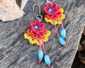 May include: Handmade floral earrings with a crochet design. The earrings feature a vibrant color palette of red, yellow, and orange, with blue beads dangling from pink threads. The earrings are set against a rustic wooden background.