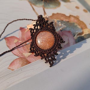 May include: A handcrafted macrame necklace featuring a round, sparkling, orange-toned gemstone pendant. The pendant is encased in a dark brown, intricately knotted macrame design. The necklace is displayed on a floral patterned surface.