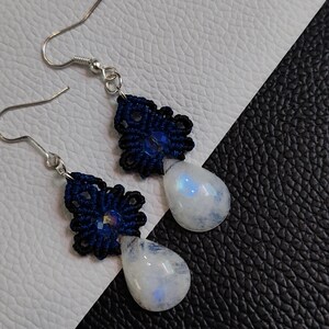 May include: A pair of handmade earrings featuring a dark blue macrame design with a teardrop-shaped, iridescent moonstone. The earrings have silver-colored ear wires and are displayed on a black and white textured surface.