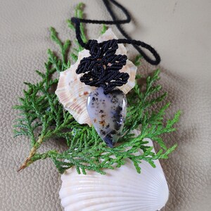 May include: A black macrame pendant with a grey and black agate stone, resting on a seashell and green foliage.