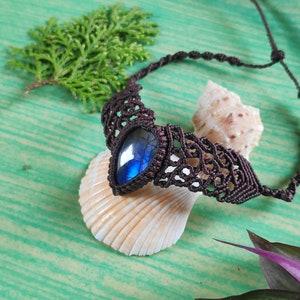 May include: A dark brown macrame necklace with a blue labradorite gemstone pendant. The necklace is displayed on a seashell and a green surface.