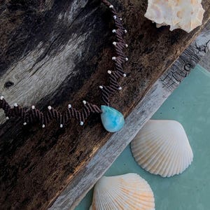 May include: A handmade macrame necklace with a teardrop-shaped, light blue pendant. The brown cord is accented with small white beads. The necklace is displayed with seashells on a weathered wooden surface, evoking a beachy, bohemian aesthetic.