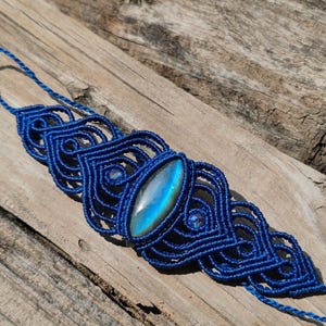 May include: A dark blue macrame bracelet featuring an elongated, iridescent labradorite gemstone at its center. The intricate knotted cord design surrounds the stone, accented by two smaller, round, faceted blue beads on either side of the center.