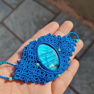 May include: A blue macrame pendant with a large oval-shaped labradorite gemstone in the center. The pendant is accented with small green beads.