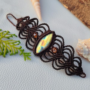 May include: A dark brown macrame bracelet featuring an elongated marquise-shaped labradorite gemstone with iridescent yellow and blue flashes, accented by two small round amber-coloured beads set within intricate knotted cord patterns.