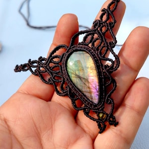 May include: A teardrop-shaped labradorite gemstone pendant set in dark brown macrame cord. The stone displays iridescent flashes of green, yellow, and purple. The intricate knotted cordwork forms a triangular frame around the central stone.