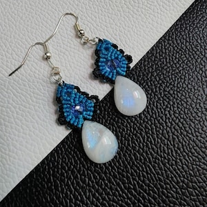 May include: A pair of handmade earrings featuring teardrop-shaped moonstone beads. The earrings have a macrame design in shades of blue and black, with silver-colored ear wires. The earrings are displayed on a black and white textured surface.