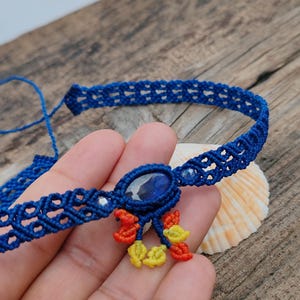 May include: A handmade blue macrame choker necklace with a central oval-shaped pendant. The pendant features a blue stone and is adorned with small orange and yellow accents. The necklace is displayed on a wooden surface.