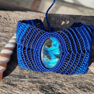 May include: A handcrafted macrame bracelet in shades of blue and black, featuring an oval labradorite stone at the center. The bracelet is made with intricate knotting and has a woven appearance. The labradorite stone displays iridescent blue and green hues.