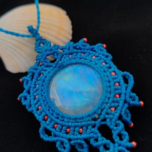 May include: A handmade macrame necklace with a large, round, iridescent blue gemstone pendant. The pendant is surrounded by intricate turquoise macrame detailing, accented with small red beads. The necklace is displayed against a black background with a seashell.