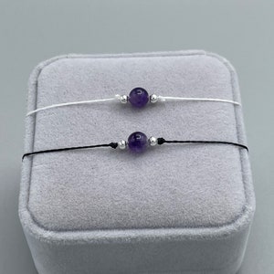 Könnte beinhalten: Zwei zarte Armbänder mit einer einzelnen Amethyst-Edelsteinperle und silbernen Akzenten. Ein Armband hat ein weißes Band und das andere ein schwarzes Band. Die Armbänder werden auf einer grauen Samtbox präsentiert.