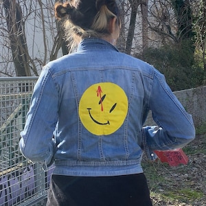 Könnte beinhalten: Eine Person trägt eine blaue Jeansjacke mit einem gelben Smiley-Gesicht mit einem roten Strich durch auf dem Rücken.