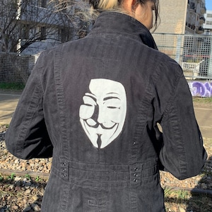 Könnte beinhalten: Schwarze Jacke mit einem weißen Grafikdruck des Anonymous-Masks auf der Rückseite.