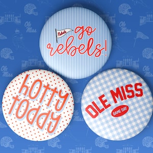 May include: Three circular pinback buttons featuring Ole Miss themes. The top button has blue stripes and says go rebels! with a flag icon. The bottom left button has red polka dots and says hotty toddy. The bottom right button has blue gingham.